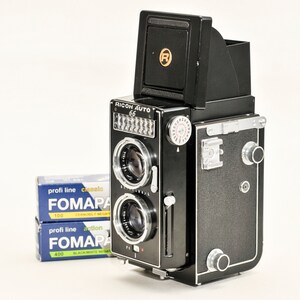 May include: A black Ricoh Auto 66 twin-lens reflex camera with two lenses and a viewfinder. The camera has a silver dial and a flash mount. Two boxes of Fomapan film are next to the camera. The camera has the Ricoh logo in gold.