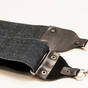 Puede incluir: Una correa de cámara de mezclilla azul oscuro con detalles de cuero negro y cierres metálicos. La correa está hecha de tela de mezclilla azul oscuro y presenta extremos de cuero negro asegurados con remaches metálicos. Cierres plateados.