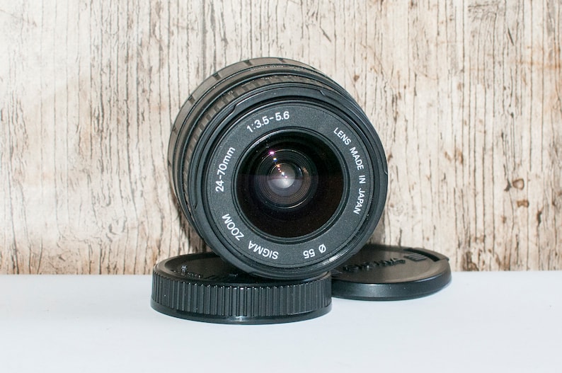 May include: A black Sigma 24-70mm zoom lens with a 1:3.5-5.6 aperture. The lens is made in Japan and has a black lens cap.