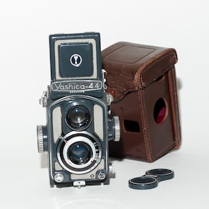 May include: A vintage Yashica-44 twin-lens reflex camera with a dark gray body and silver accents. The camera is accompanied by a brown leather case and a lens cap. The Yashica-44 is a classic film camera.