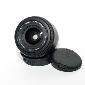 May include: A black Sigma Super-Wide II camera lens with a 52mm diameter, f=24mm, and a lens cap. The lens is multi-coated and made in Japan. The lens cap is black and has the Sigma logo.