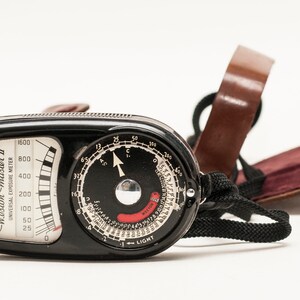 May include: A black Weston Master II universal exposure meter with a silver dial and a red needle. The meter has a black leather strap and a brown leather case.