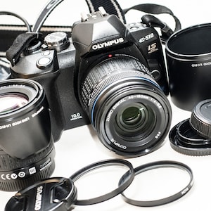 Puede incluir: Una cámara digital Olympus E-510 negra con múltiples lentes, tapas de lentes y un parasol. La cámara tiene un objetivo zoom adjunto, con el texto "OLYMPUS DIGITAL 40-150mm 1:4-5.6". También son visibles accesorios adicionales.
