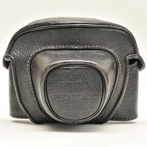 May include: A black leather camera case with white stitching. The case has a curved top and a rounded front, with the brand name "ASAHI PENTAX" embossed on the front. The case is designed to protect a camera.