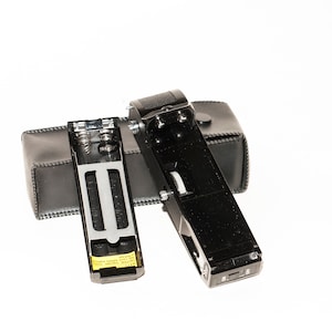 Puede incluir: Flash de cámara vintage negro con una funda de cuero. El flash está abierto, revelando el compartimento de la batería. La funda es rectangular y negra. El flash tiene una parte superior texturizada y un dial lateral.