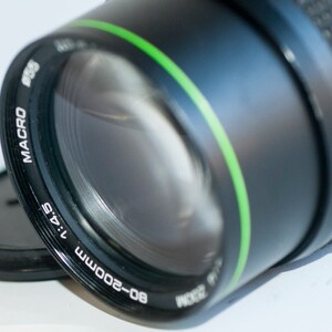 Op de afbeelding: Een zwarte camera lens met een groene streep rond de rand. De lens is gemarkeerd met "80-200mm 1:4.5 MACRO".