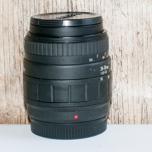 May include: A black camera lens with a 24-70mm zoom range and a 1:3.5-5.6 aperture. The lens has a red dot on the bottom.
