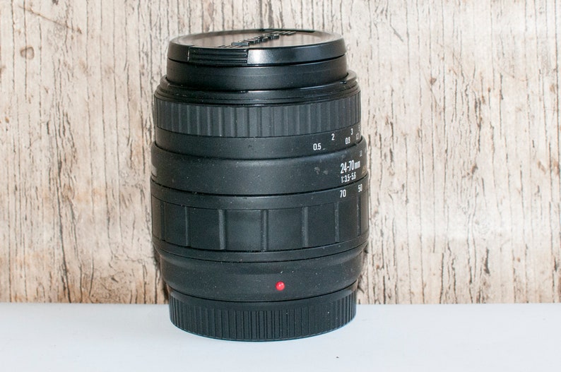May include: A black camera lens with a 24-70mm zoom range and a 1:3.5-5.6 aperture. The lens has a red dot on the bottom.