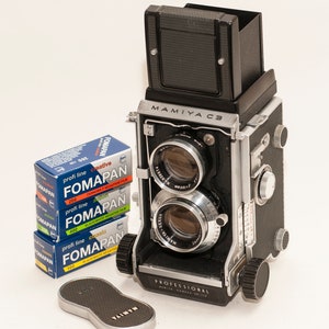 May include: A black and silver Mamiya C3 twin-lens reflex camera with a top-mounted viewfinder, two stacked lenses, and side adjustment knobs. Beside the camera are three blue and white boxes of Fomapan film and a separate black Mamiya lens cap.