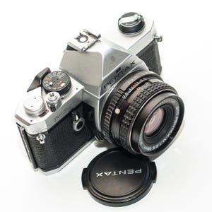 Puede incluir: Una cámara de película Pentax MX plateada y negra con una lente negra y una tapa de lente. La tapa de la lente está en la parte inferior derecha y la cámara está en ángulo hacia el espectador. La cámara tiene un agarre de cuero negro y varios diales y botones.