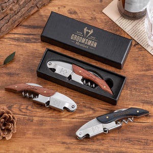 Pode incluir: Três abridores de garrafas de vinho com cabos de madeira e componentes de aço inoxidável são exibidos em uma superfície de madeira. Um abridor está em uma caixa preta com o texto "Groomsman" e o nome "Heather". Um pinha e um copo também são visíveis.