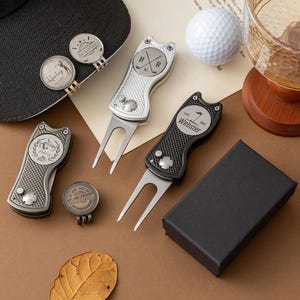Könnte beinhalten: Golfzubehör, darunter Pitchgabeln und Ballmarker, sind auf einer braunen Oberfläche angeordnet. Die Pitchgabeln sind silber und schwarz, mit personalisiertem Text. Eine schwarze Geschenkbox ist ebenfalls vorhanden.