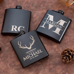 Könnte beinhalten: Drei schwarze Flachmänner mit silbernen Gravuren. Einer hat die Initialen "RC", ein anderer den Namen "MARGAN" in einem großen Buchstaben "M", und der dritte ein Hirschgeweih-Design mit dem Namen "MICHAEL" und dem Datum "20-12-2024".