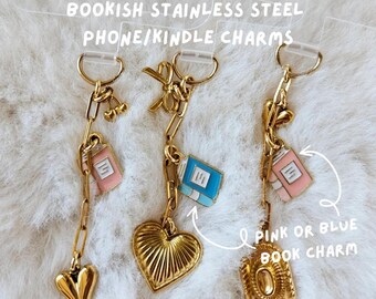 Op de afbeelding: Goudkleurige roestvrijstalen telefoon-/Kindle-bedels. De bedels hebben een roze of blauwe boekenbedel, een hart en een gouden ketting. De tekst op de afbeelding luidt "BOOKISH STAINLESS STEEL PHONE/KINDLE CHARMS" en "PINK OR BLUE BOOK CHARM."