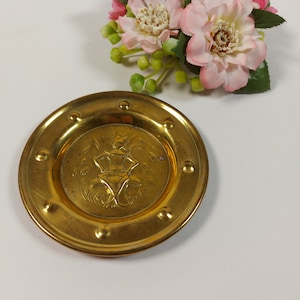 May include: A small, round, gold-coloured metal dish with a raised design of a figure in a dress. The dish has a beaded edge and is on a white background.
