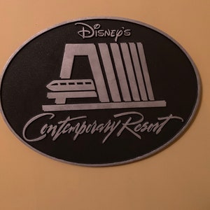 Pu&ograve; includere: Un cartello ovale nero e argento con il testo "Disney's Contemporary Resort" e un'immagine stilizzata della monorotaia del resort.