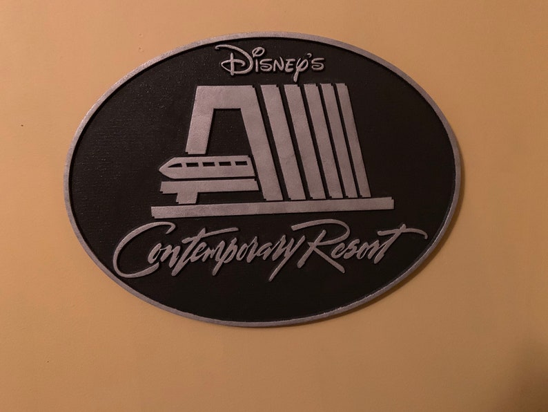 Pu&ograve; includere: Un cartello ovale nero e argento con il testo "Disney's Contemporary Resort" e un'immagine stilizzata della monorotaia del resort.