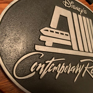 Pu&ograve; includere: Una placca rotonda nera e argentata con il testo "Disney's Contemporary Resort" e un'immagine stilizzata del sistema di monorotaia del resort.