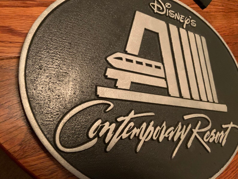 Pu&ograve; includere: Una placca rotonda nera e argentata con il testo "Disney's Contemporary Resort" e un'immagine stilizzata del sistema di monorotaia del resort.