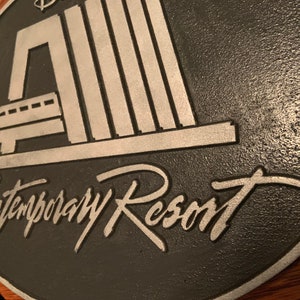 Pu&ograve; includere: Una placca in metallo nero e argento con il testo "Contemporary Resort" e un'immagine stilizzata di una monorotaia che entra in un edificio.
