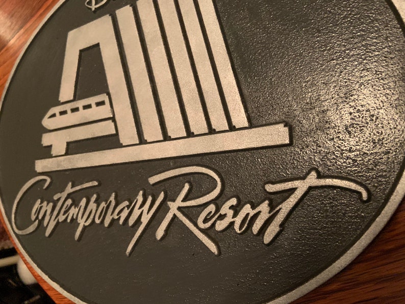 Pu&ograve; includere: Una placca in metallo nero e argento con il testo "Contemporary Resort" e un'immagine stilizzata di una monorotaia che entra in un edificio.