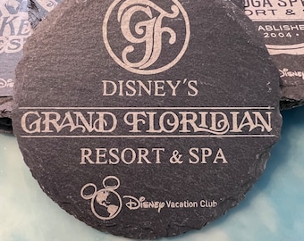 May include: A black slate coaster with the Disney's Grand Floridian Resort & Spa logo. The logo features a stylized "GF" monogram and the text "Disney's Grand Floridian Resort & Spa". The coaster also features the Disney Vacation Club logo.
