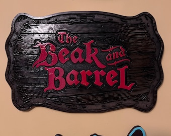 May include: A rectangular wooden sign with a dark brown finish and a scalloped edge, featuring the text The Beak and Barrel in raised, bold red lettering. The wood grain texture is visible across the surface of the sign, which is mounted on a wall.