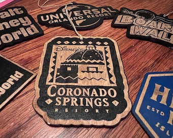 May include: Wooden laser-cut ornaments featuring various resort logos, including "Coronado Springs Resort" and "Universal Orlando Resort." The ornaments are brown with black detailing and have a rustic aesthetic. Some have string for hanging.