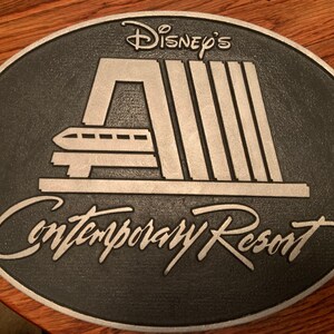 Pu&ograve; includere: Una placca in metallo argentato con sfondo nero. La placca presenta un'immagine stilizzata della stazione del monorail del Contemporary Resort con il testo "Disney's Contemporary Resort".