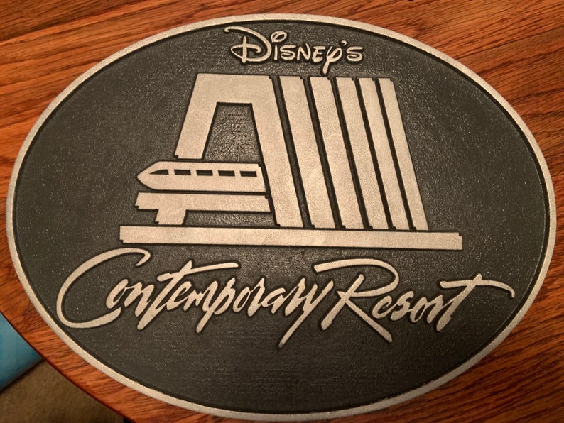 Pu&ograve; includere: Una placca in metallo argentato con sfondo nero. La placca presenta un'immagine stilizzata della stazione del monorail del Contemporary Resort con il testo "Disney's Contemporary Resort".