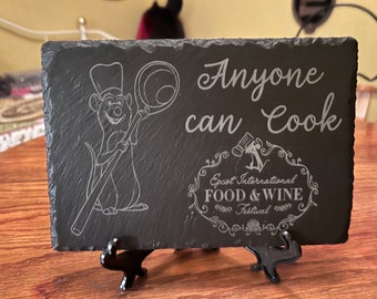 May include: Black slate sign with a white engraved image of a chef rat holding a whisk. The sign reads "Anyone can Cook" and "Epcot International Food & Wine Festival".