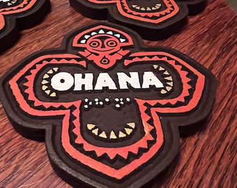 May include: Four dark brown, flower-shaped coasters with the word "OHANA" in white. The coasters have red and gold tribal-style designs. The coasters are on a wooden surface.