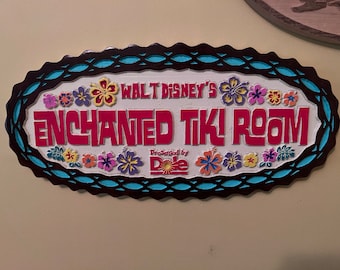 Può includere: Cartello in legno per l'Enchanted Tiki Room, con la scritta "Walt Disney's Enchanted Tiki Room" in rosso. Il cartello è decorato con fiori colorati e un bordo turchese e marrone.
