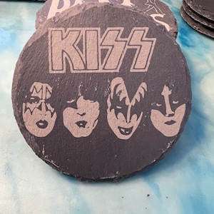 May include: Black slate coaster with the word "KISS" printed in white and four white silhouettes of the band members' faces.
