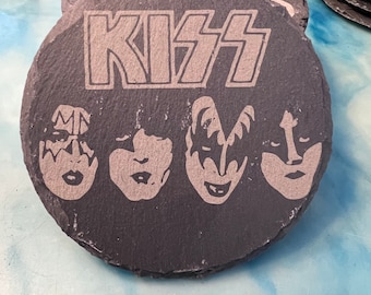 May include: A stack of dark gray slate coasters. The top coaster features the band name "KISS" in large, bold letters, with the faces of the band members below. Additional coasters with a similar design are visible in the background.