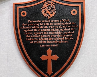 May include: Shield-shaped wall plaque with a black background and orange trim. The plaque features a lion's head, a cross, and text from Ephesians 6:11-12. The shield is approximately 10 inches tall and 8 inches wide.