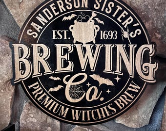 May include: Round wooden sign with the text "Sanderson Sister's Brewing Co. Premium Witches Brew" in black and tan. The sign features bats, a cauldron, and a spiderweb design. The sign is hanging from a natural twine string.
