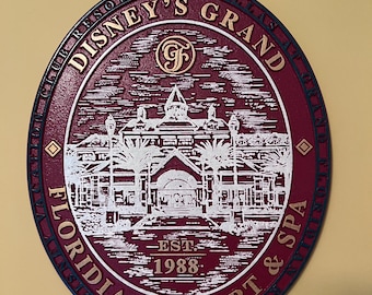 May include: A maroon and gold oval sign for Disney's Grand Floridian Resort & Spa. The sign features the resort's name, a detailed illustration of the building, and the establishment year, 1988. The sign has a dark blue border.
