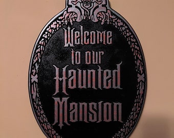 May include: A black and silver oval sign with a decorative frame and the text "Welcome to our Haunted Mansion".