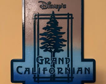 May include: A rectangular sign with the text "Disney's Grand Californian Hotel". The sign features a stylized tree design within a rectangular frame, with a gradient blue and brown color scheme.
