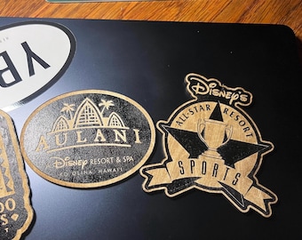 May include: Wooden Disney resort and spa themed decorative plaques. One plaque is oval-shaped with the text "AULANI" and "Disney Resort & Spa". The other plaque is star-shaped with the text "Disney's All-Star Resort SPORTS".