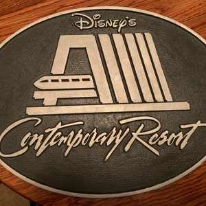 Pu&ograve; includere: Una placca in metallo nero e argento con il testo "Disney's Contemporary Resort" e un'immagine stilizzata del sistema di monorotaia del resort.