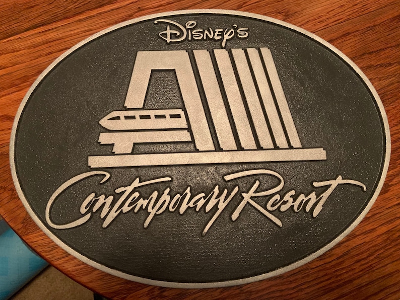 Pu&ograve; includere: Una placca in metallo nero e argento con il testo "Disney's Contemporary Resort" e un'immagine stilizzata del sistema di monorotaia del resort.