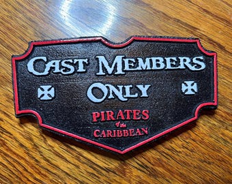 May include: Black sign with red trim and the words "CAST MEMBERS ONLY" in white. Below, it reads "PIRATES of the CARIBBEAN" in red. The sign has a textured surface and decorative corners.
