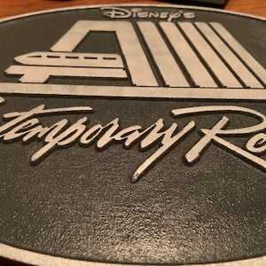 Pu&ograve; includere: Una placca in metallo nero e argento con il testo "Disney's Contemporary Resort" in un carattere stilizzato. La placca presenta una silhouette argentata della stazione monorotaia iconica del resort.