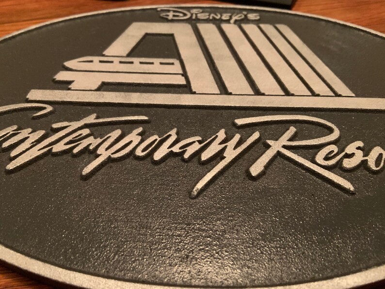 Pu&ograve; includere: Una placca in metallo nero e argento con il testo "Disney's Contemporary Resort" in un carattere stilizzato. La placca presenta una silhouette argentata della stazione monorotaia iconica del resort.