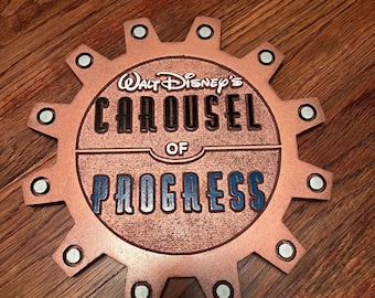 May include: A copper-colored gear-shaped wall sign featuring the text Walt Disney's CAROUSEL OF PROGRESS. The sign has a textured metallic finish with raised black lettering for CAROUSEL and raised blue lettering for PROGRESS, accented by silver rivets.
