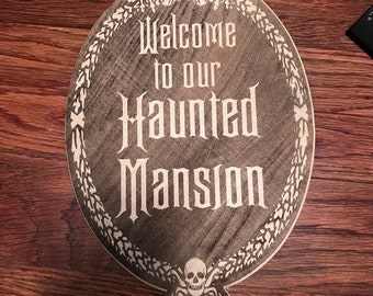 May include: A wooden sign with a distressed gray finish and white lettering. The sign reads "Welcome to our Haunted Mansion". The sign is decorated with a skull and crossbones at the bottom and a gargoyle at the top.