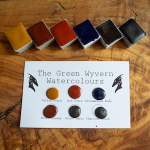 Puede incluir: Un conjunto de pinturas de acuarela en varios colores, incluyendo ocre amarillo, ocre rojo y azul ultramar, dispuestas sobre una superficie de madera. El conjunto incluye una tarjeta con el texto "The Green Wyvern Watercolours".