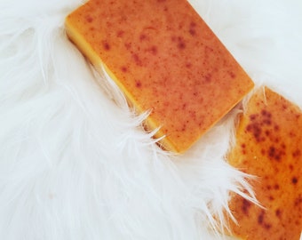 May include: Two bars of orange-coloured soap on a white fluffy surface. The soap has a speckled appearance.